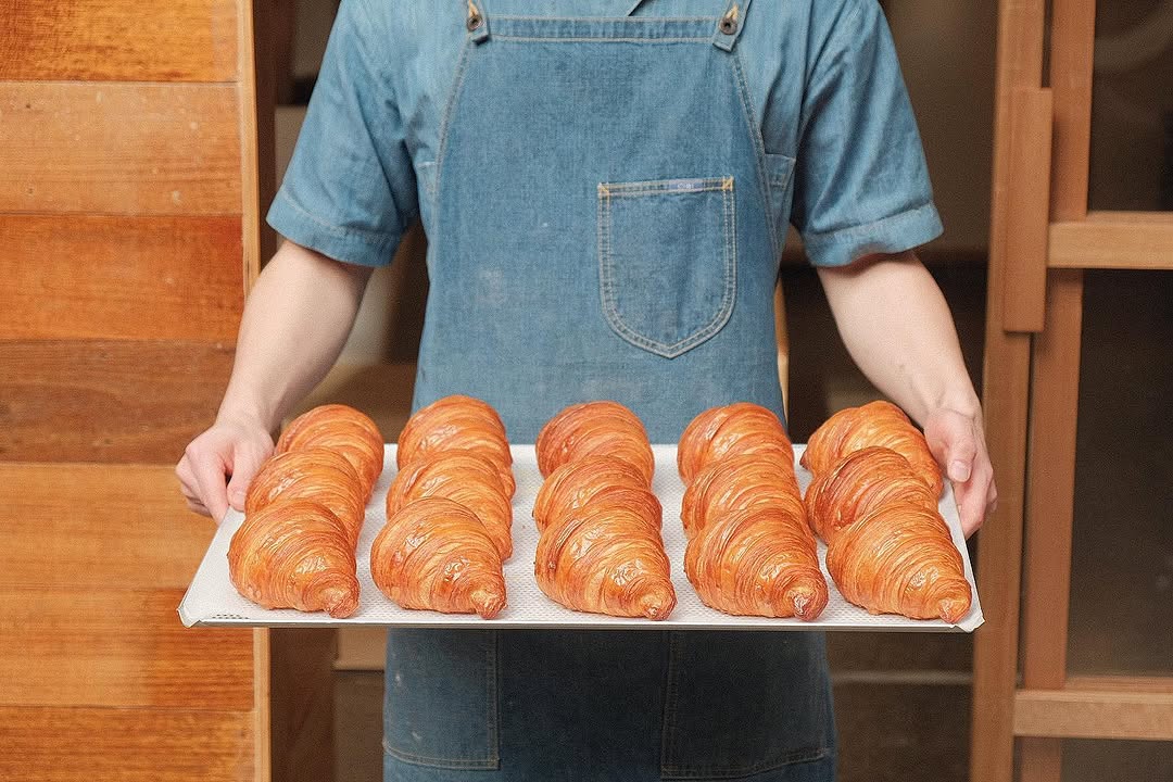 A baker in a denim apron carrying a tray of glossy fresh croissants
