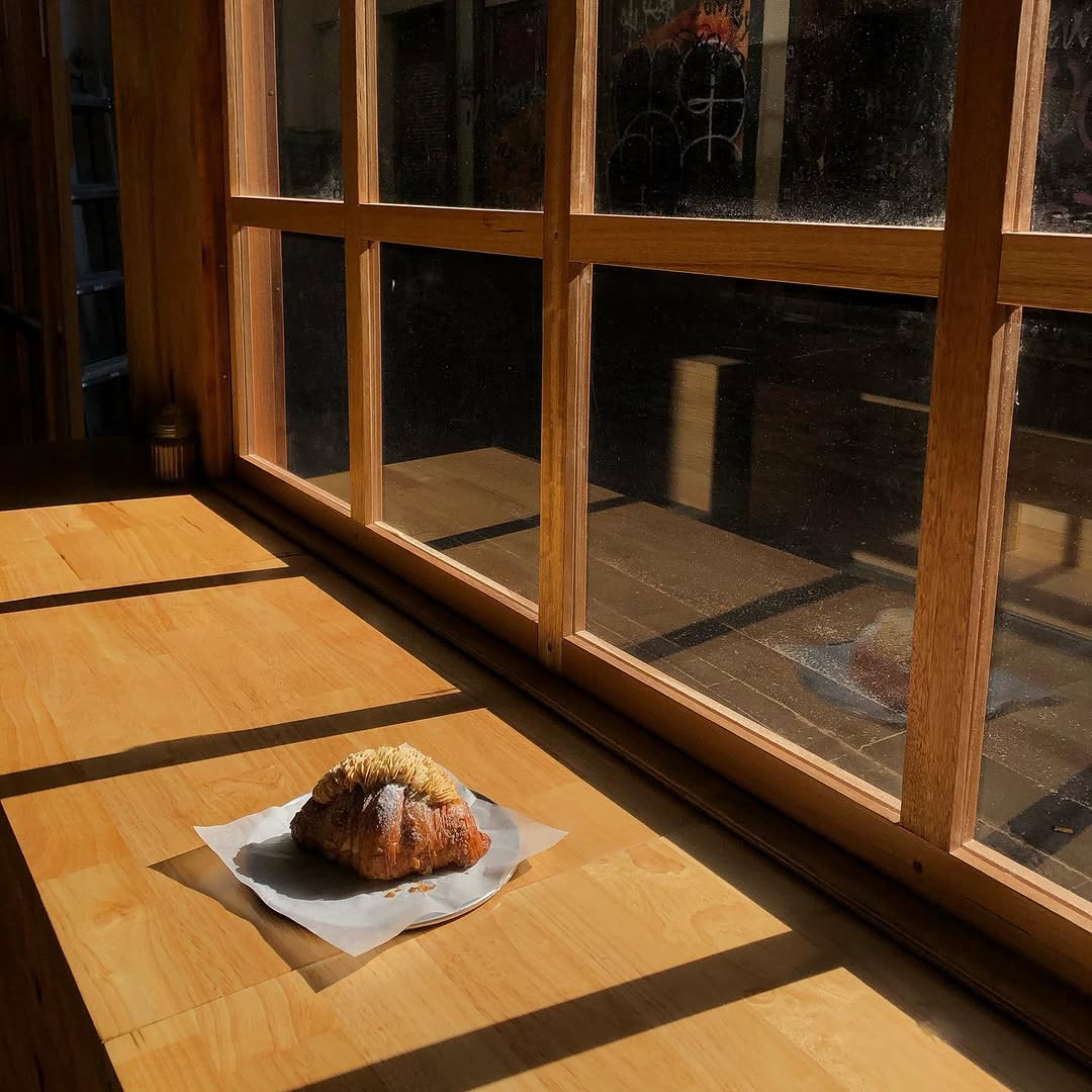 A single croissant on parchment, lit by a grid of morning sun through timber windows