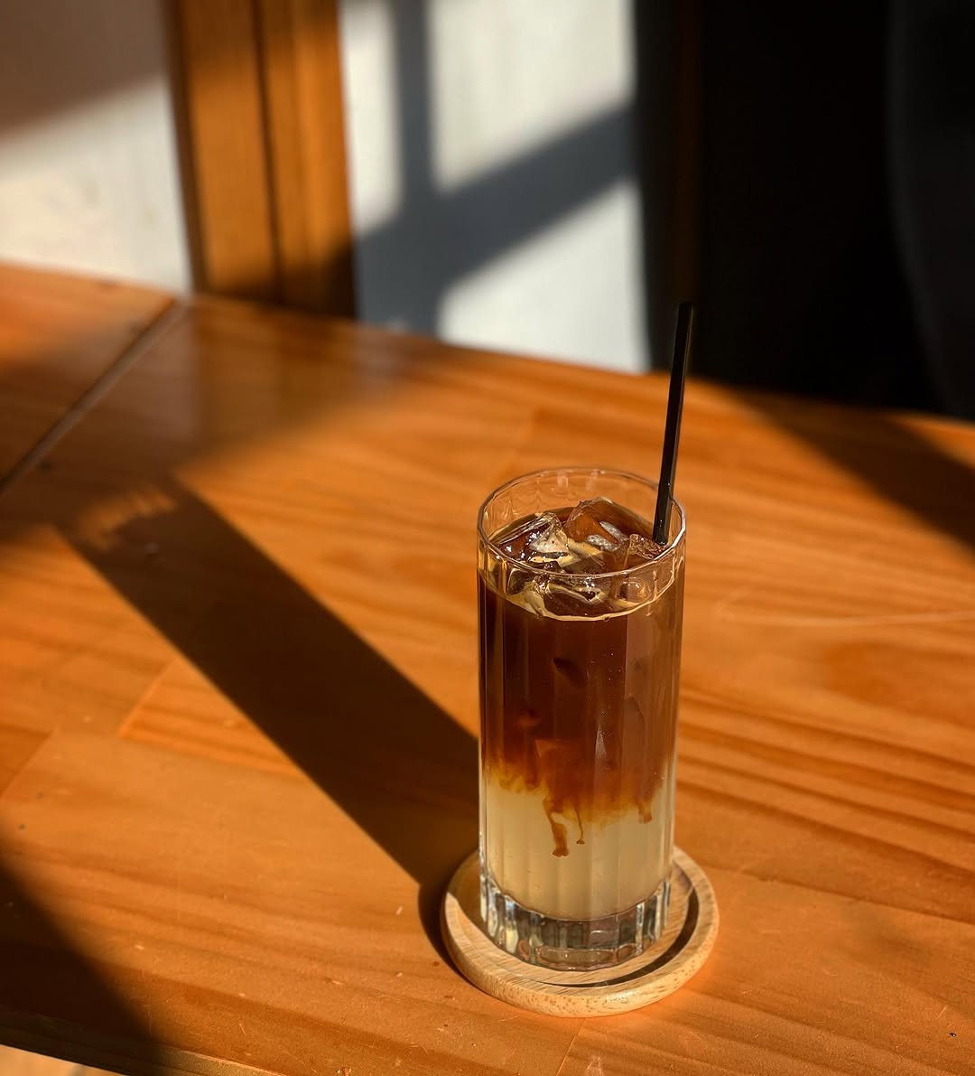 A tall glass of iced hojicha latte on a wooden coaster in warm morning light